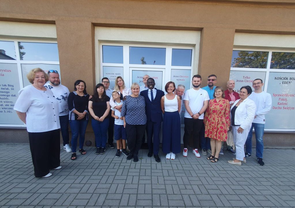 Kim Jesteśmy 10 Grupa ludzi pozująca do zdjęcia przed budynkiem z napisem 'Kościół Boży Eden'. Uczestnicy stoją w dwóch rzędach, uśmiechając się do kamery. W tle widoczne są okna z napisami, takimi jak 'Jezus Chrystus jest Panem' oraz 'Jezus Chrystus wyzwala'. Atmosfera jest radosna i pełna wspólnoty.