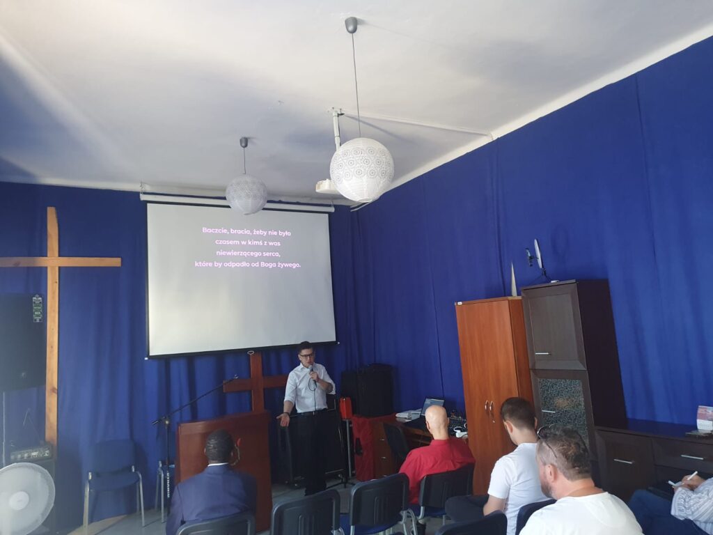 Kim Jesteśmy 1 Mężczyzna przemawiający do grupy ludzi w sali z niebieskimi zasłonami i dużym drewnianym krzyżem. Na ekranie wyświetlony jest tekst biblijny, a uczestnicy siedzą w skupieniu, słuchając mowy.