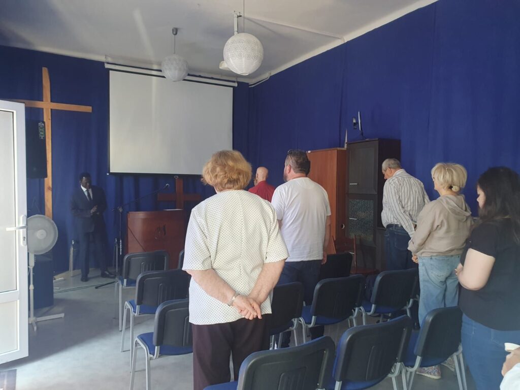 Kim Jesteśmy 9 Grupa osób stojących w sali z niebieskimi zasłonami, zwrócona w kierunku podium i ekranu. Mężczyzna w garniturze stoi przy podium z mikrofonem, obok znajduje się duży drewniany krzyż. Uczestnicy mają ręce złożone za plecami, w atmosferze skupienia i modlitwy.