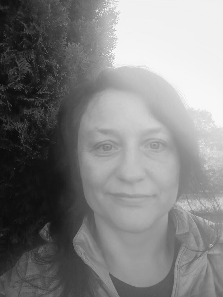 Czarno-białe zdjęcie kobiety z długimi włosami, patrzącej prosto w kamerę. W tle widać drzewo iglaste oraz delikatnie rozmytą scenerię na zewnątrz.