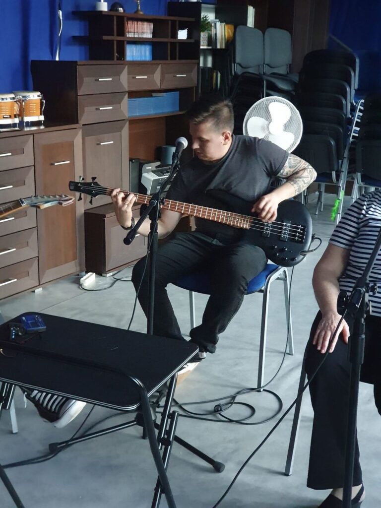 Kim Jesteśmy 7 Mężczyzna gra na czarnej gitarze basowej podczas próby muzycznej. Przed nim ustawiony jest mikrofon. W tle znajdują się drewniane meble oraz wentylator, a na szafkach widać bębny perkusyjne.