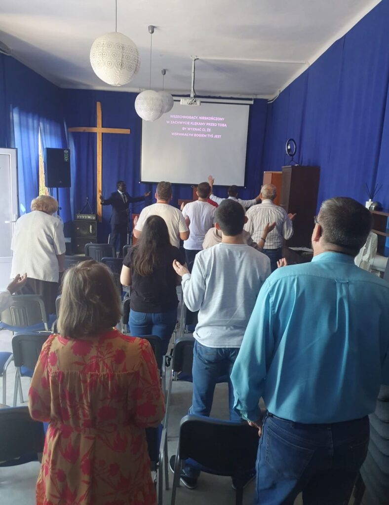 Kim Jesteśmy 5 Grupa osób stojąca w sali podczas nabożeństwa, z rękami uniesionymi w geście uwielbienia. Na ścianie w tle znajduje się drewniany krzyż, a na ekranie wyświetlają się słowa pieśni: 'Wszechmogący, nieskończony, w zachwycie klękamy przed Tobą...'. Atmosfera jest uroczysta, a sala udekorowana jest niebieskimi zasłonami.