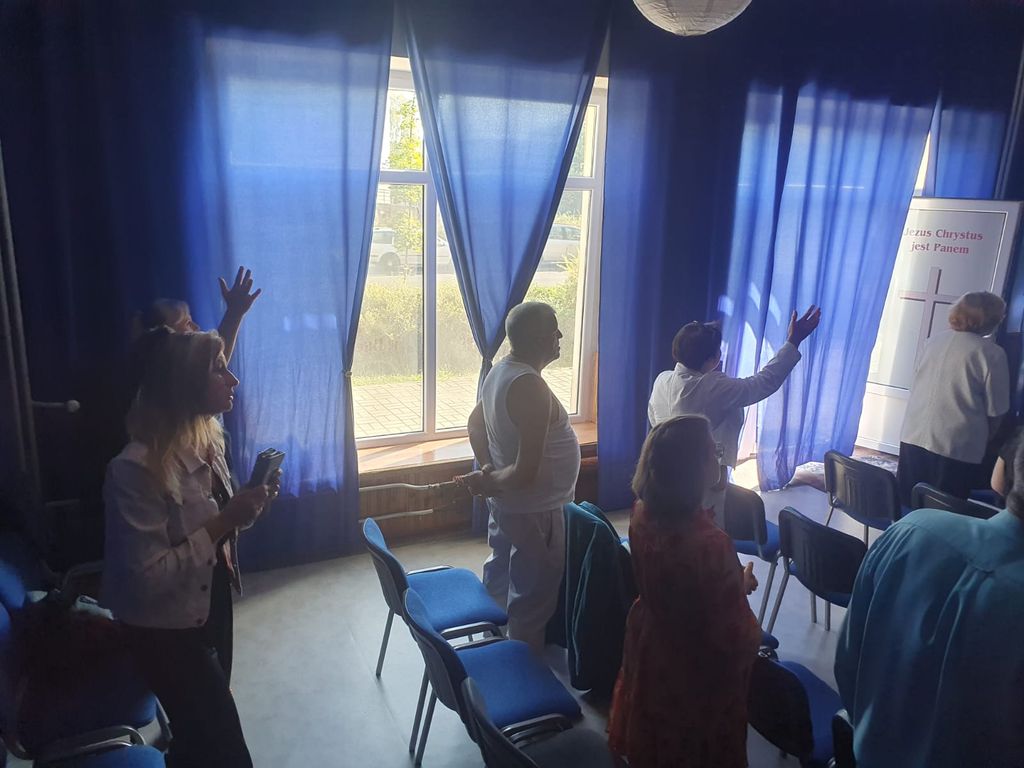 Kim Jesteśmy 4 Grupa osób stojących przed oknem z niebieskimi zasłonami, unoszących ręce w geście modlitwy lub uwielbienia. W tle widać napis 'Jezus Chrystus jest Panem' obok krzyża. Sala jest jasno oświetlona światłem dziennym wpadającym przez okno.