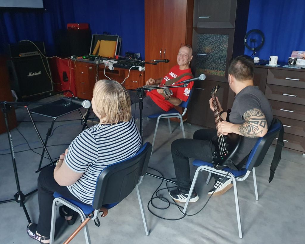 Kim Jesteśmy 8 Trio muzyków podczas próby. Kobieta siedzi przy mikrofonie, zwrócona tyłem do kamery. Mężczyzna w czerwonej koszulce z napisem 'Zaufaj Jezusowi' gra na gitarze elektrycznej, uśmiechając się, a mężczyzna z tatuażem na ramieniu gra na gitarze basowej. W tle widać sprzęt muzyczny i wzmacniacze.