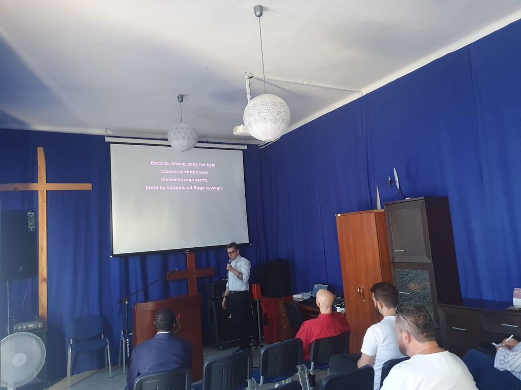 Mężczyzna przemawiający do niewielkiej grupy osób w sali z niebieskimi zasłonami i drewnianym krzyżem. Na ekranie wyświetlany jest tekst biblijny: 'Baczcie, bracia, żeby nie było czasem w kimś z was niewierzącego serca, które by odpadło od Boga żywego'. Uczestnicy słuchają siedząc na krzesłach, a atmosfera jest spokojna i refleksyjna.