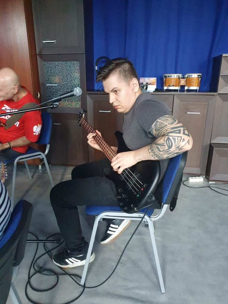 Kim Jesteśmy 3 Mężczyzna z tatuażem na ramieniu gra na czarnej gitarze basowej podczas spotkania muzycznego. Obok niego, po lewej stronie, widać mężczyznę w czerwonej koszulce z mikrofonem. W tle znajdują się drewniane meble oraz instrumenty perkusyjne.