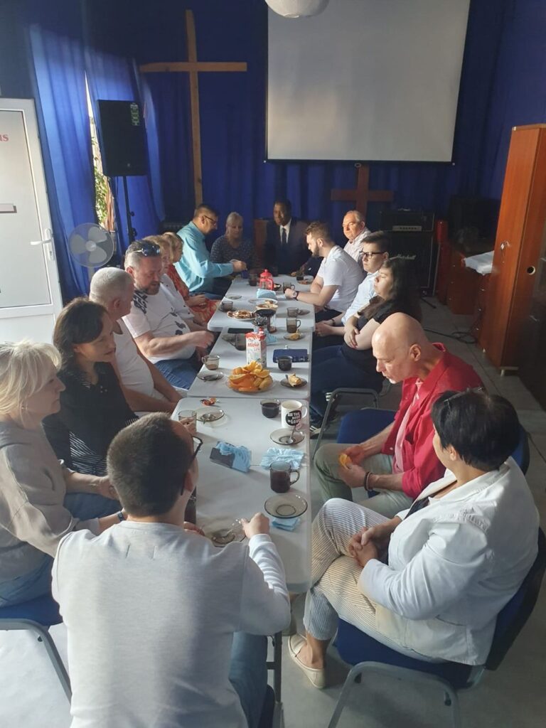 Kim Jesteśmy 2 Grupa osób siedząca przy długim stole podczas spotkania towarzyskiego w jasnej sali. Na stole znajdują się filiżanki, napoje oraz owoce, w tym pomarańcze. Uczestnicy prowadzą rozmowy w luźnej atmosferze. W tle widać niebieskie zasłony oraz drewniany krzyż na ścianie.